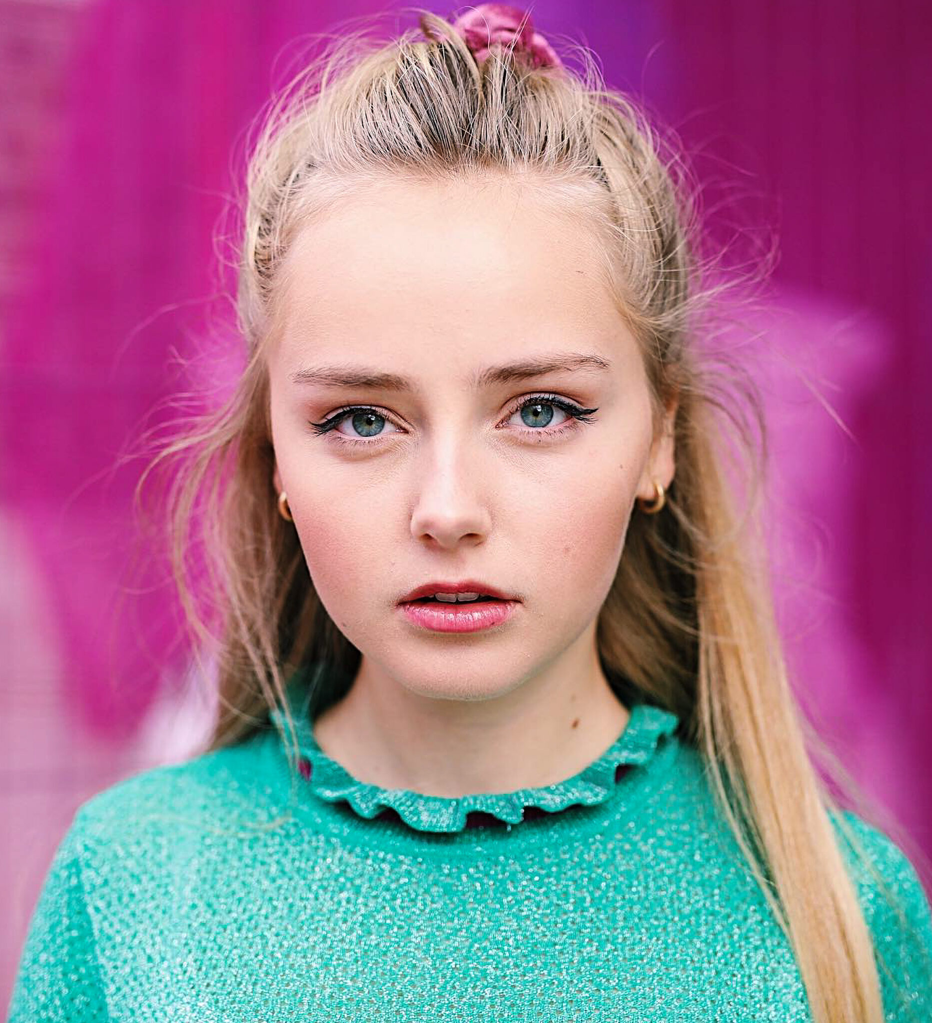 A young, blonde-haired woman wearing a green top with a ruffled collar, with a neutral facial expression.