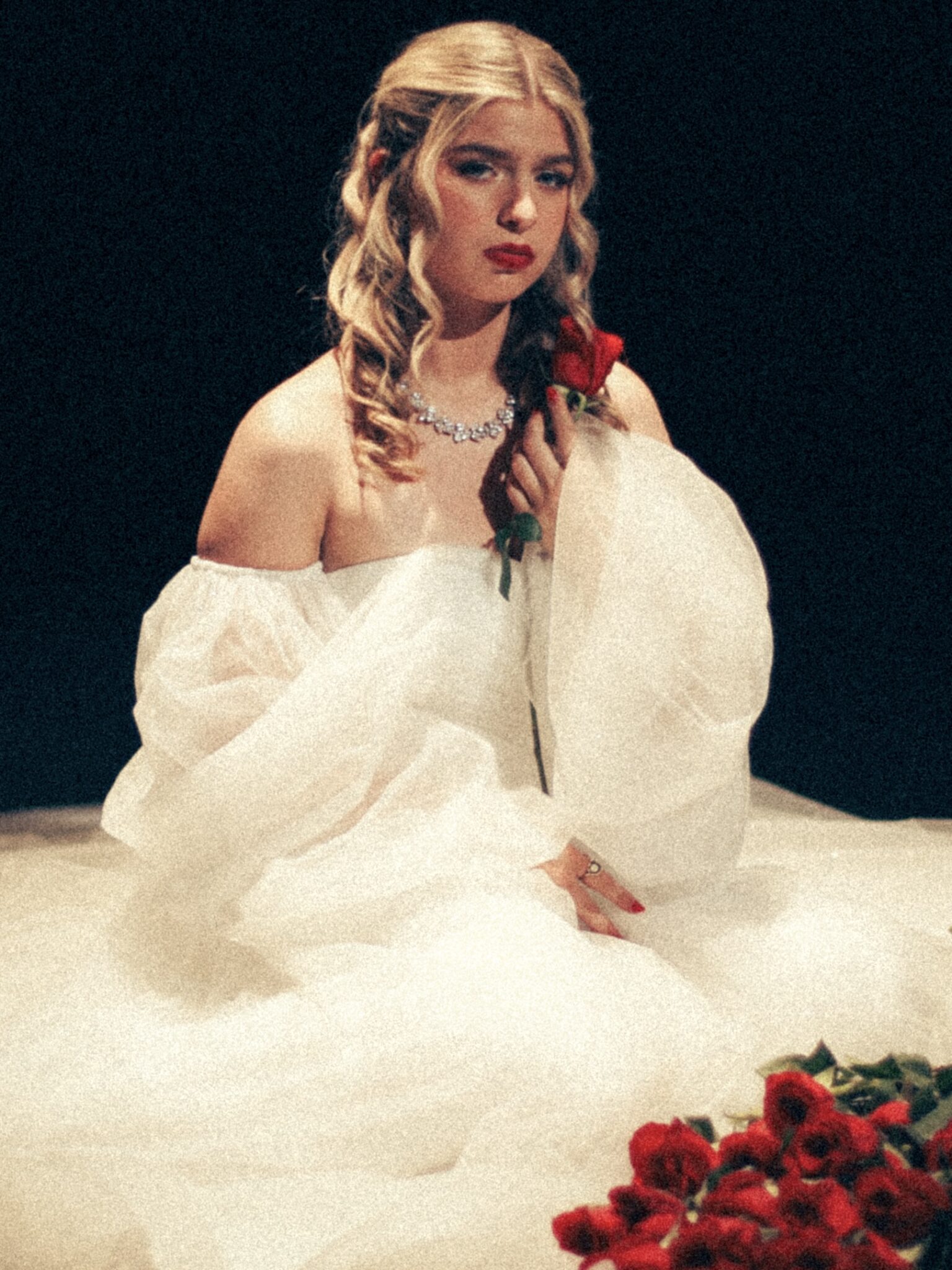 A young and blonde woman with a somber look wearing a ruffled, off-the shoulder dress with a rose in her hand and a bouquet of roses in front of her.
